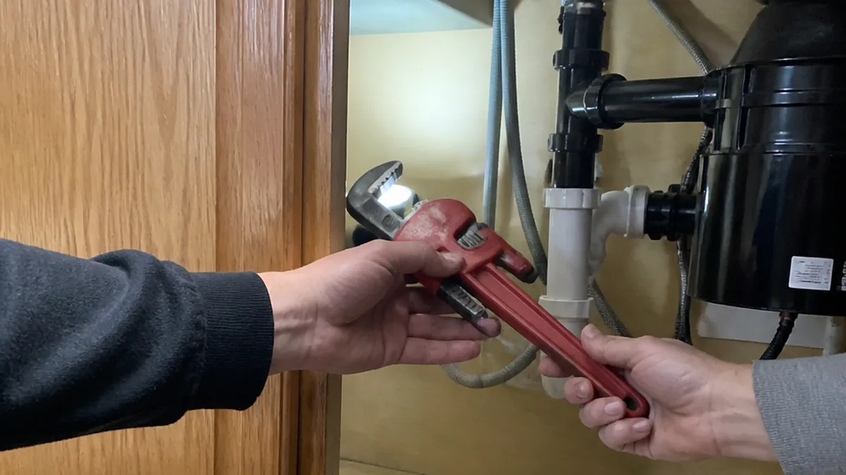 A young plumber's apprentice handing a pipe wrench to a senior plumber under a sink