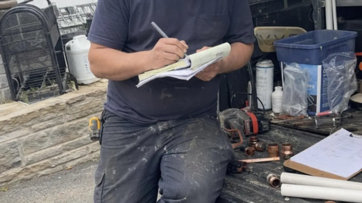 A plumber at a kitchen table reviewing a flat-rate pricing book on a clipboard